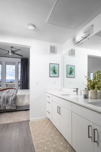 A bathroom with a large mirror, a bed with a grey comforter, and a fan.